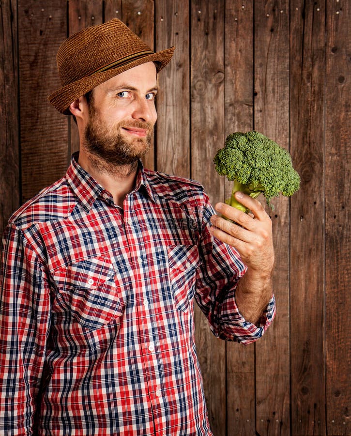 Happy smiling caucasian forty years old farmer or gardener in a hat holding broccoli in hand on rustic vintage planked wood background - agriculture. Food production - vegetables. Happy smiling caucasian forty years old farmer or gardener in a hat holding broccoli in hand on rustic vintage planked wood background - agriculture. Food production - vegetables.