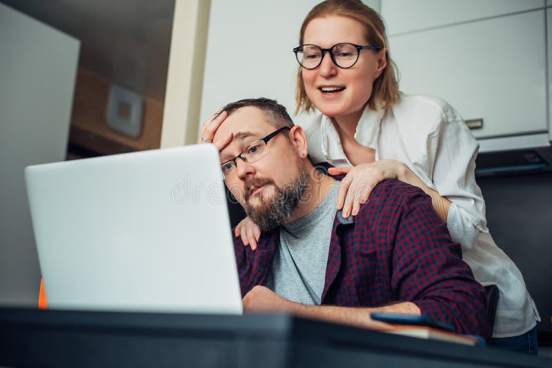 Brodaty mężczyzna i kobieta z okularami za pomocą laptopa w domu komunikują się ze znajomymi przez Internet śmiechem i grimace zdjęcia stock