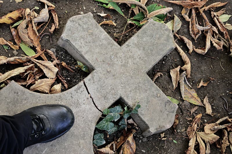 Broken stone cemetery cross lying on ground with black leather boot standing on it