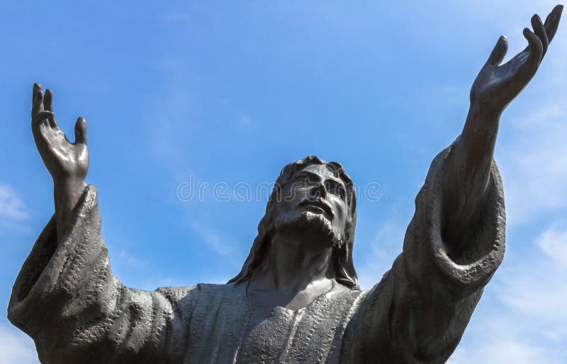 Bronze statue of Jesus looking at the sky with both arms up. On background a blue sky whit white clouds