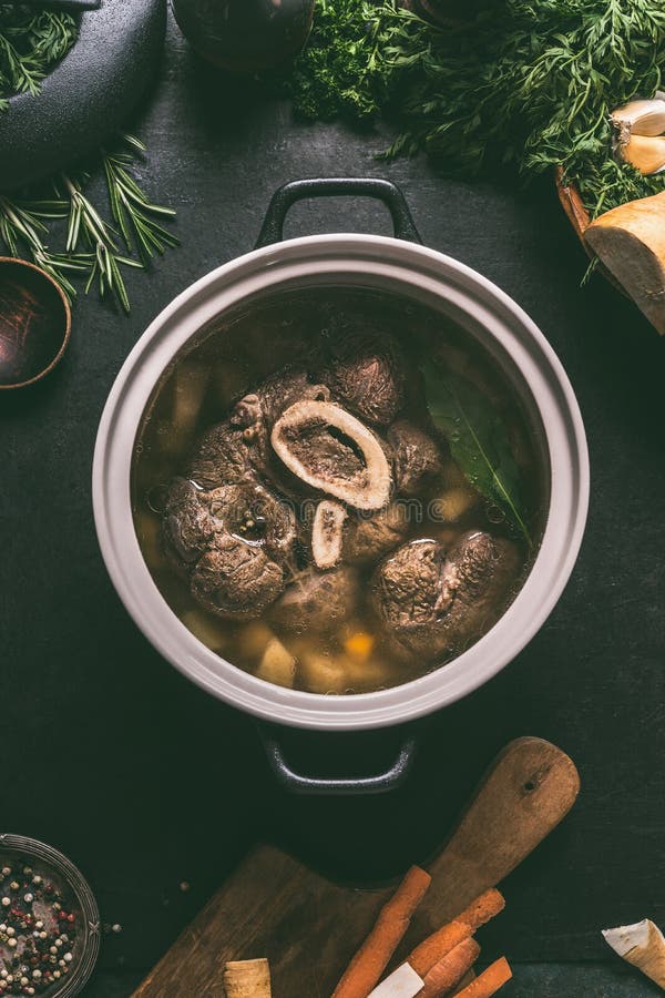 Broth of beef meat shin with bone in cooking pot on dark kitchen table background with ingredients for soup, top view. Meat broth