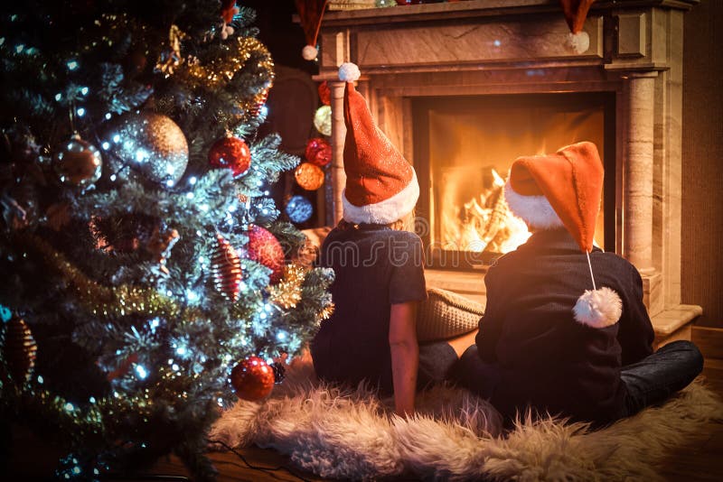 Back view, brother and sister wearing Santa`s hats warming next to a fireplace in a living room decorated for Christmas.