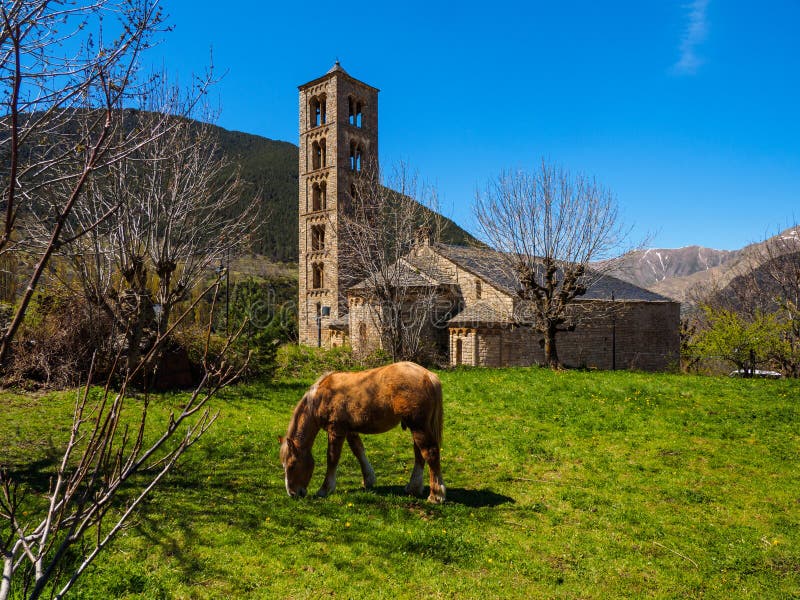 Brown horse grazing in the green grass at the foot of the Romanesque church of Taull, with the bell tower and the abside in the foreground. Montaña del Pirieno, Vall de Boi, Unesco Heritage, Catalonia, Spain. Brown horse grazing in the green grass at the foot of the Romanesque church of Taull, with the bell tower and the abside in the foreground. Montaña del Pirieno, Vall de Boi, Unesco Heritage, Catalonia, Spain