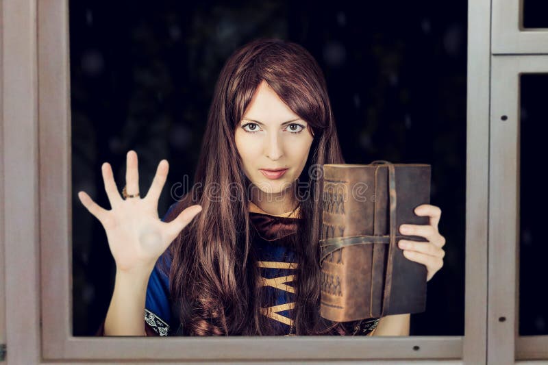 Beautiful young halloween witch wearing vintage gothic dress holding magical book of spells in old leather cover she leaned her hand against the window outside. Beautiful young halloween witch wearing vintage gothic dress holding magical book of spells in old leather cover she leaned her hand against the window outside