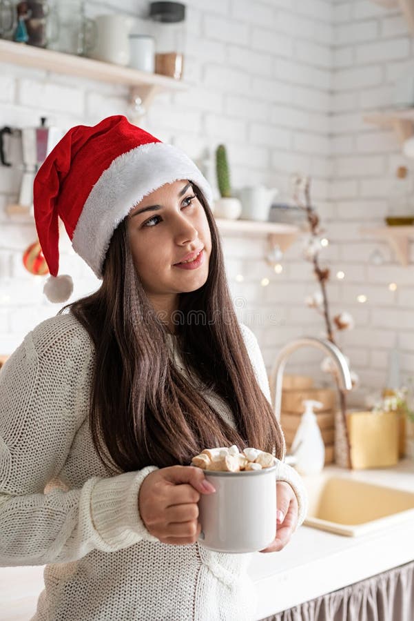 Brunetka w kapeluszu santa trzymająca filiżankę piankowego kakao w kuchni obrazy royalty free