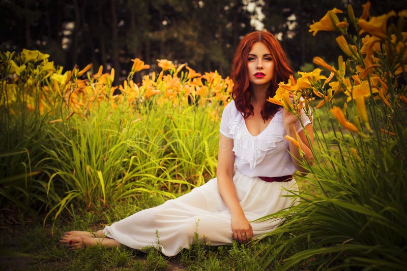 Brunette caucasian woman in white dress at the park in red and yellow flowers on a summer sunset holding flowers sitting on the gr