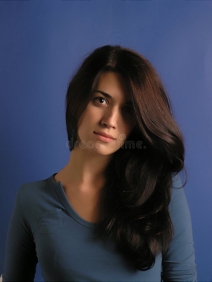 Portrait of a beautiful brunette woman, her head tilted and her long hair draped around over one shoulder. Provocative expression. Head and shoulders shot. Isolated on a blue background. Portrait of a beautiful brunette woman, her head tilted and her long hair draped around over one shoulder. Provocative expression. Head and shoulders shot. Isolated on a blue background.