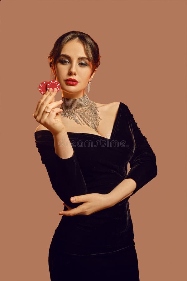 Brunette female in black dress and shiny jewelry. She is showing two red chips, posing on brown studio background. Poker
