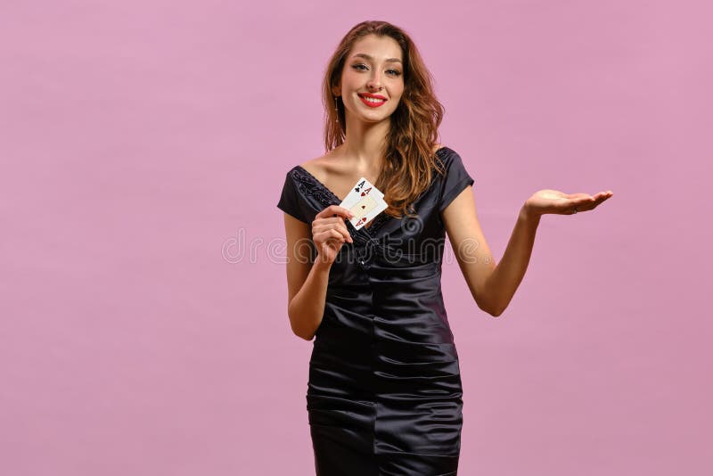 Brunette female in black dress. Showing two aces, smiling, holding something, posing on pink background. Poker, casino