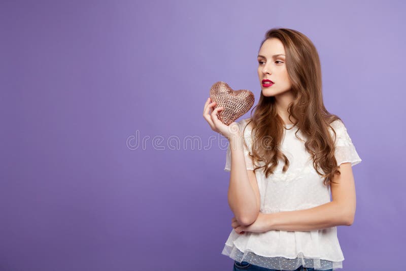 The brunette girl is holding a decorative heart in her hand. On a lilac background. concept for Valentine`s Day. place for text stock photo The brunette girl is holding a decorative heart in her hand. On a lilac background. concept for Valentine`s Day. place for text stock photo