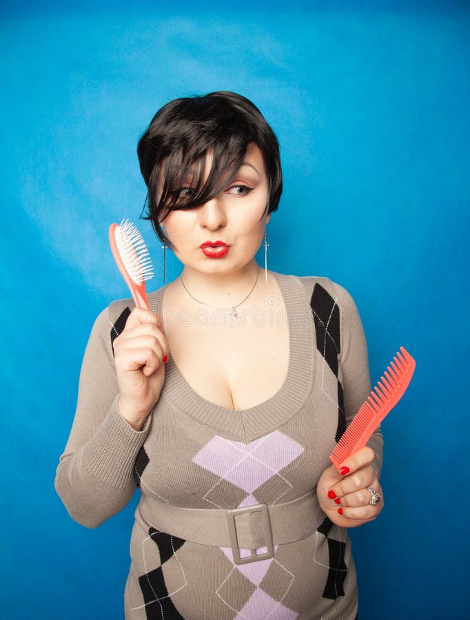 Brunette girl with short hair compares comb and brush for hair on blue studio background