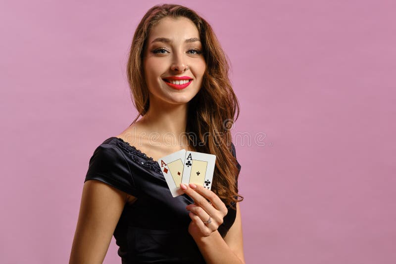 Brunette lady with earring in nose, in black dress. Showing two playing cards, smiling, posing on pink studio background