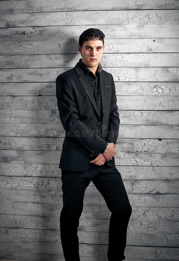 Brunette man in black suit leaning against white leaning wall.