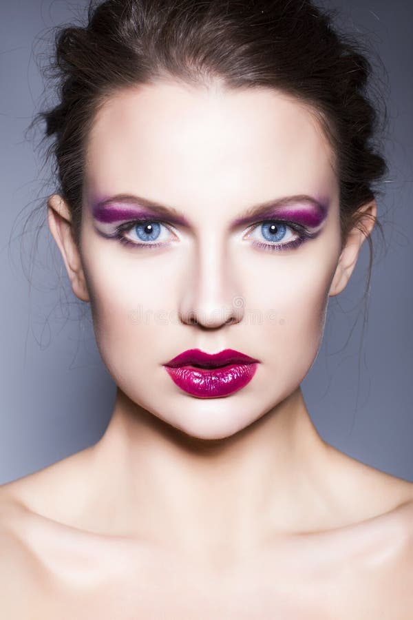 Brunette woman with creative make up violet eye shadows full red lips, blue eyes and curly hair with her hand on her face