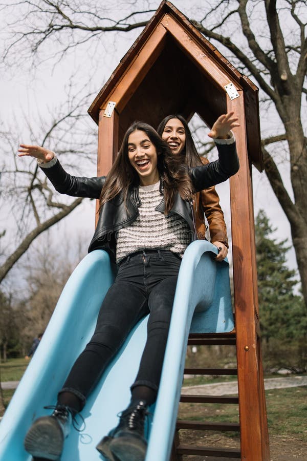 Brunette women sliding in children`s play park. Identical twins having fun on slide in playground during spring season. Brunette women sliding in children`s play park. Identical twins having fun on slide in playground during spring season.