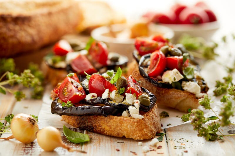 Bruschetta con la berenjena asada a la parrilla, los tomates de cereza, el queso feta, las alcaparras y las hierbas aromáticas fr fotografía de archivo Bruschetta con la berenjena asada a la parrilla, los tomates de cereza, el queso feta, las alcaparras y las hierbas aromáticas fr fotografía de archivo