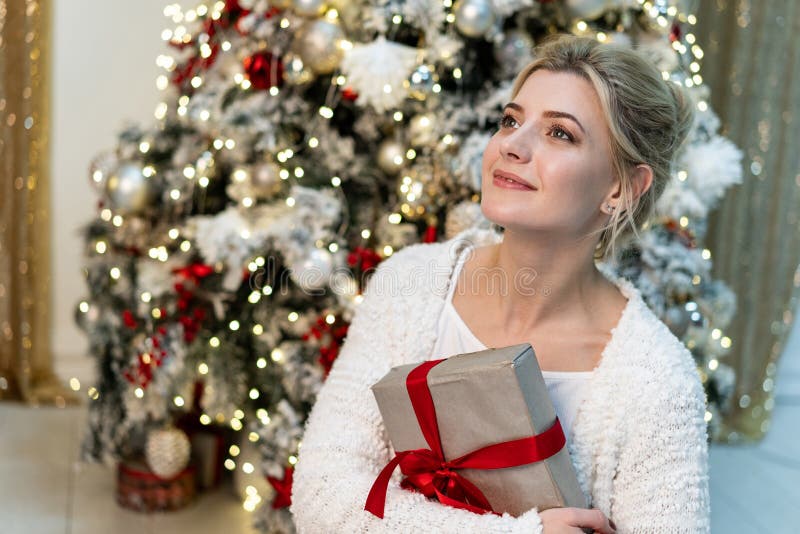 Brustbild des schönen jungen blonden Mädchens in der weißen Strickjacke, die mit Geschenk nahe dem Weihnachtsbaum aufwirft lizenzfreie stockfotos Brustbild des schönen jungen blonden Mädchens in der weißen Strickjacke, die mit Geschenk nahe dem Weihnachtsbaum aufwirft lizenzfreie stockfotos