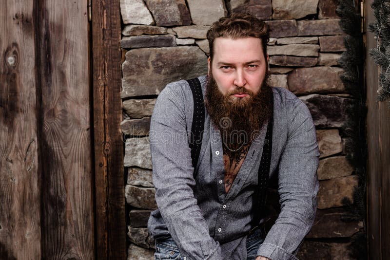Brutale sterke mens met een baard en tatoegeringen op zijn handen gekleed in modieuze vrijetijdskledingstribunes op de achtergron royalty-vrije stock fotografie Brutale sterke mens met een baard en tatoegeringen op zijn handen gekleed in modieuze vrijetijdskledingstribunes op de achtergron royalty-vrije stock fotografie