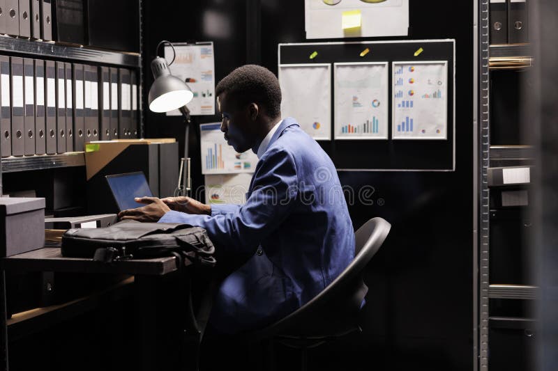 Bookkeeper reading administrative report, organizing management documents in storage room. African american businessman wearing formal suit working late at night in corporate depository. Bookkeeper reading administrative report, organizing management documents in storage room. African american businessman wearing formal suit working late at night in corporate depository