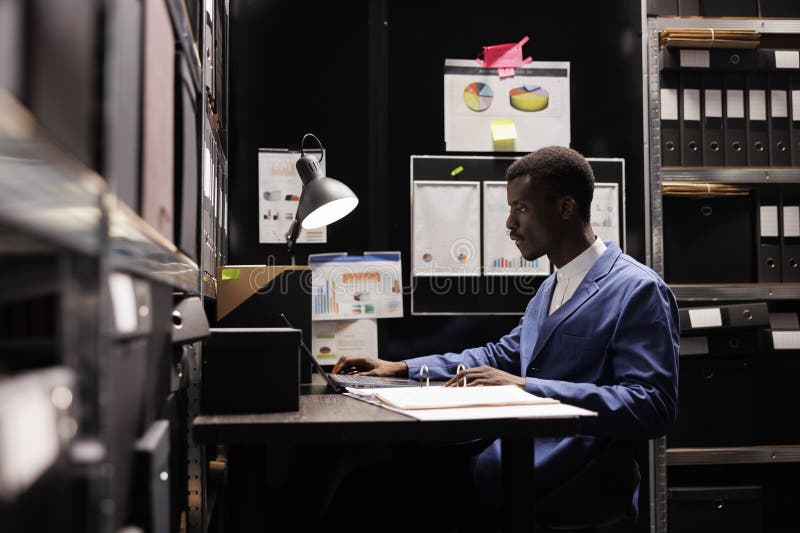 African american bookkeeper checking administrative files on laptop computer, working overtime in corporate depository. Businessman analyzing bureaucracy report in storage room. African american bookkeeper checking administrative files on laptop computer, working overtime in corporate depository. Businessman analyzing bureaucracy report in storage room