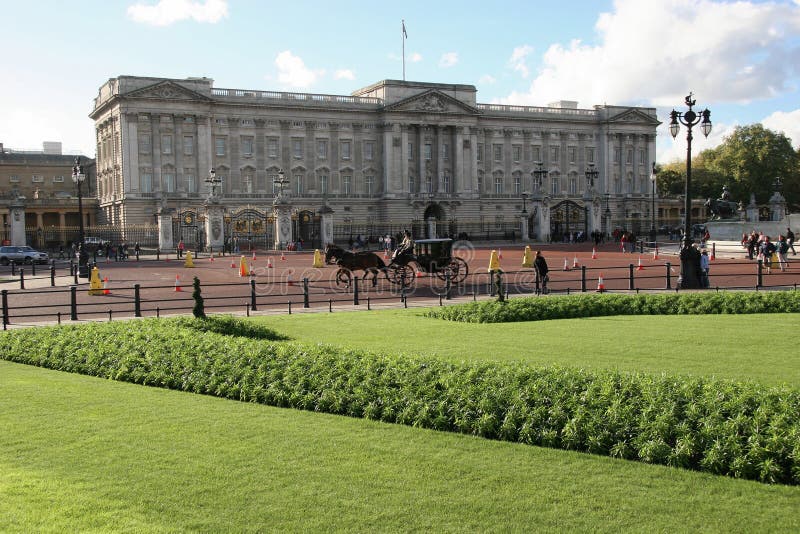 Full frontal view of Buckingham Palace over green grass and manicured flower bed. No cars. Horse drawn Brougham carriage being ridden in front of the gates. Very few people. Blue sky. Royal Standard flying. Full frontal view of Buckingham Palace over green grass and manicured flower bed. No cars. Horse drawn Brougham carriage being ridden in front of the gates. Very few people. Blue sky. Royal Standard flying.