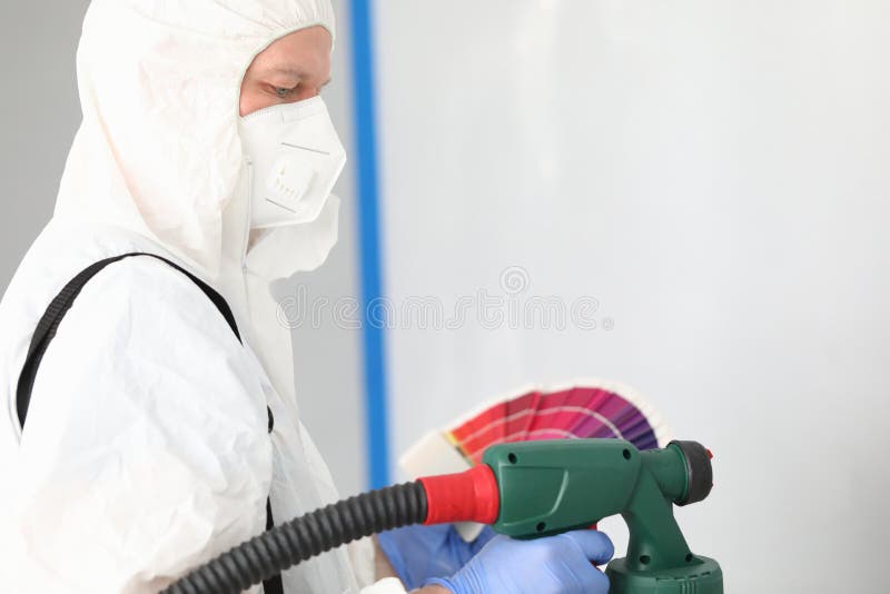 Builder in uniform chooses color for painting wall. Surface treated wall. Finishing putty to eliminate minor defects. Man is wearing protective clothing and respirator. Use electric spray gun