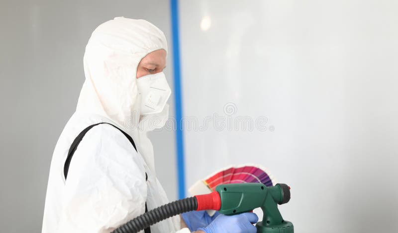 Builder in uniform chooses color for painting wall. Surface treated wall. Finishing putty to eliminate minor defects. Man is wearing protective clothing and respirator. Use electric spray gun