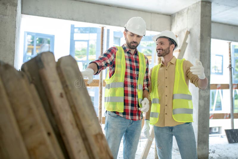 Building workers in yellow vests and gloves standing near pile of boards, discussing what to do with them, smiling stock images