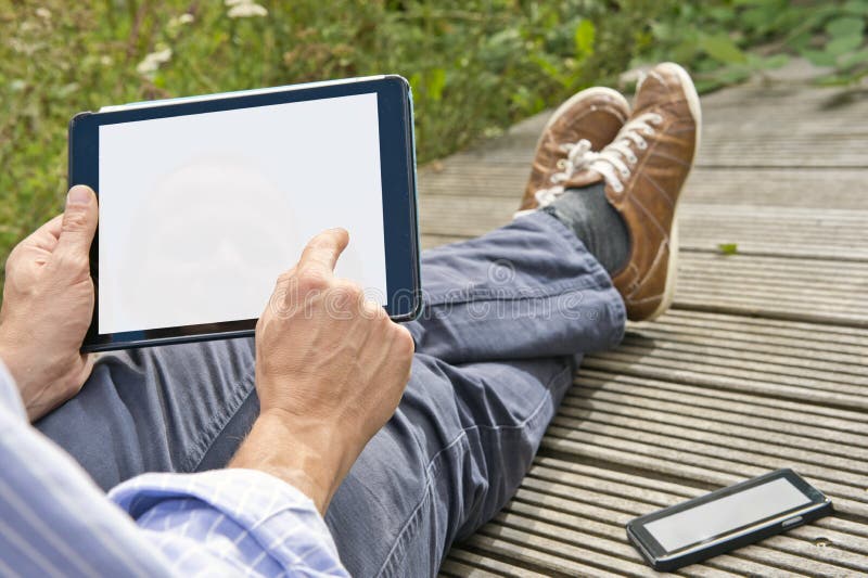 Man, tapping on a tablet, playing a game outdoors, sitting on a plank bridge, whith his phone next to him. Both can be used as presentation for mock-up apps, website and presentations. Man, tapping on a tablet, playing a game outdoors, sitting on a plank bridge, whith his phone next to him. Both can be used as presentation for mock-up apps, website and presentations.