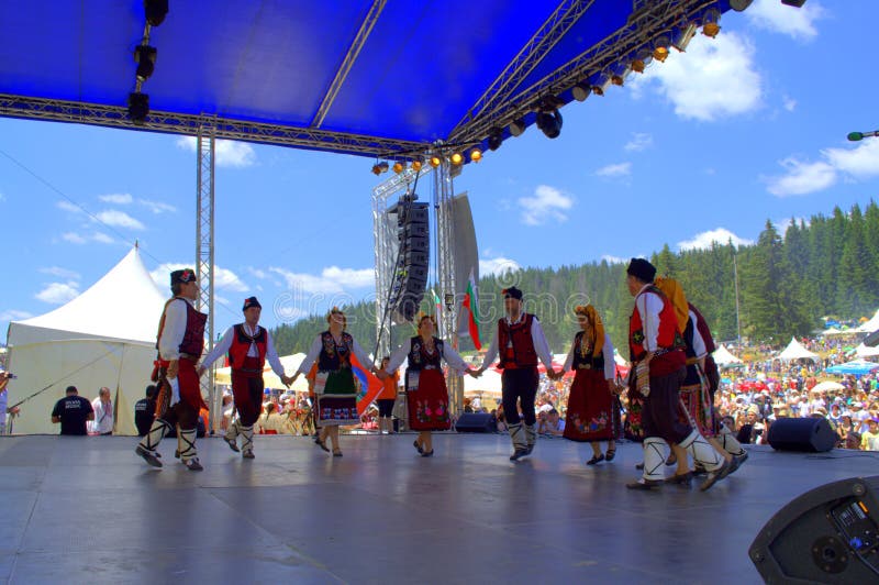 Bulgarian folklore group dressed in traditional authentic costumes dancing at scene of Folklore Festival Rozhen in beautiful scenery of Rhodope Mountains.Every 4th year in July, Bulgarians of all over the country gather below the peak of Rozhen for a festival, where singers and instrumentalists sing and play on a several scenes. Bulgarian folklore group dressed in traditional authentic costumes dancing at scene of Folklore Festival Rozhen in beautiful scenery of Rhodope Mountains.Every 4th year in July, Bulgarians of all over the country gather below the peak of Rozhen for a festival, where singers and instrumentalists sing and play on a several scenes.
