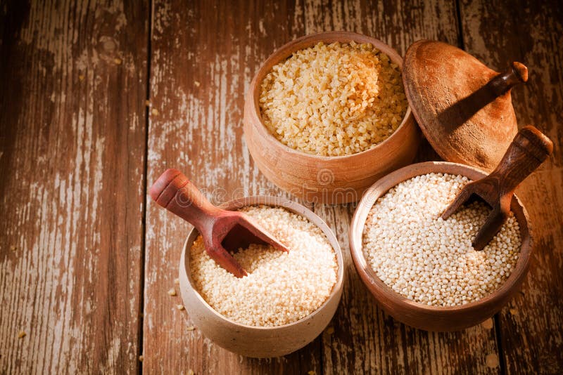 High angle of three rustic pots on a wooden background containing dried bulgur and couscous made from crushed durum and semolina wheat and quinoa seeds, a high-protein staple of the goosefoot family. High angle of three rustic pots on a wooden background containing dried bulgur and couscous made from crushed durum and semolina wheat and quinoa seeds, a high-protein staple of the goosefoot family