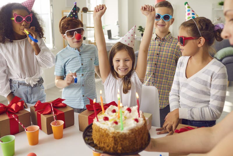 Group of happy diverse children in funny sunglasses celebrating their friend`s birthday. Mum giving cake with candles to her daughter surrounded by multiethnic classmates who are blowing party horns. Group of happy diverse children in funny sunglasses celebrating their friend`s birthday. Mum giving cake with candles to her daughter surrounded by multiethnic classmates who are blowing party horns