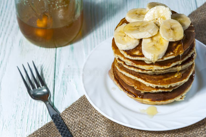 Bunt av hemlagade pannkakor med bananskivor och honung på den vita plattan med gaffel- och linneservetten på träbakgrund arkivbild Bunt av hemlagade pannkakor med bananskivor och honung på den vita plattan med gaffel- och linneservetten på träbakgrund arkivbild