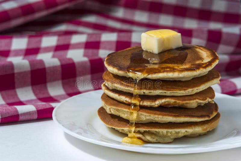 Bunt av hemlagade pannkakor med smör och honung på den vita plattan med den rutiga servetten royaltyfri bild Bunt av hemlagade pannkakor med smör och honung på den vita plattan med den rutiga servetten royaltyfri bild