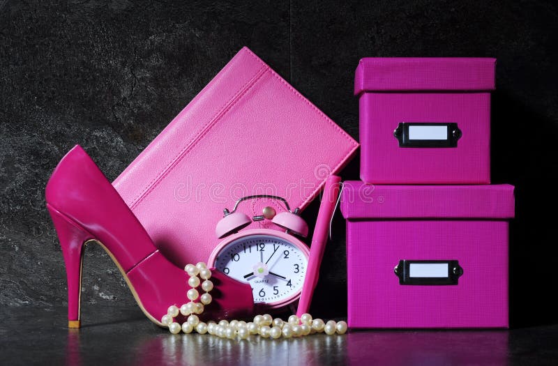 Ladies pretty pink female office desk with high heel shoe, clock, pen, stationery filing boses, pearls and diary organiser against a dramatic black background. Ladies pretty pink female office desk with high heel shoe, clock, pen, stationery filing boses, pearls and diary organiser against a dramatic black background.