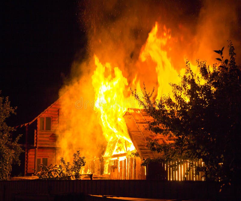 Burning wooden house at night. Bright orange flames and dense smoke from under the tiled roof on dark sky, trees silhouettes and royalty free stock images Burning wooden house at night. Bright orange flames and dense smoke from under the tiled roof on dark sky, trees silhouettes and royalty free stock images