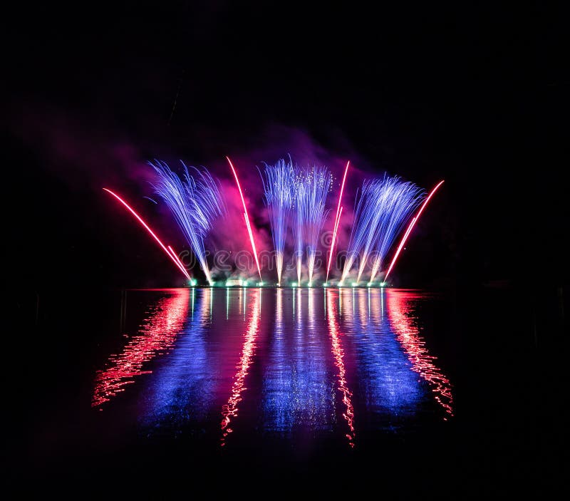 Bursts of blue and red fire in rich fireworks over Brno`s Dam with lake reflection