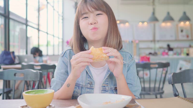 Business freelance Asian woman eating bread and drinking warm cup of coffee while sitting on table in cafe.