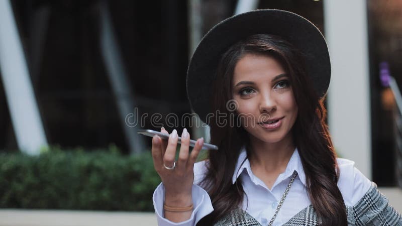 Business lady in black hat sending audio voice message on cellphone at outdoor talking to mobile assistant. Girl using