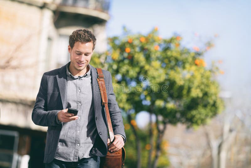 Business man using mobile phone walking in city street commuting to work with blazer and messenger bag texting on