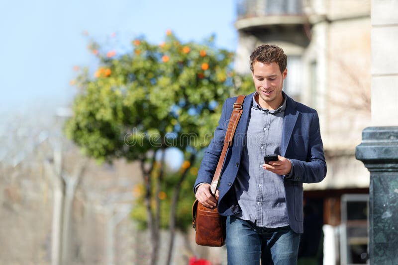 Young urban businessman professional on smartphone walking in street using mobile phone app texting sms message on smartphone wearing smart casual jacket. City lifestyle commute person walking. Young urban businessman professional on smartphone walking in street using mobile phone app texting sms message on smartphone wearing smart casual jacket. City lifestyle commute person walking.