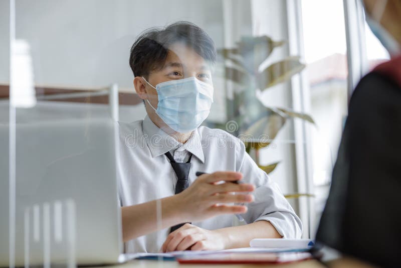 Business man workers wearing face mask and clear shield having discussion through glass partition at the office