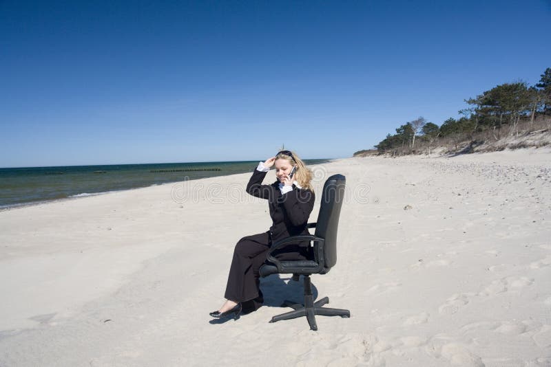 Blonde woman in business suit sitting in swivel office chair on sandy beach talking on cell phone. Blonde woman in business suit sitting in swivel office chair on sandy beach talking on cell phone