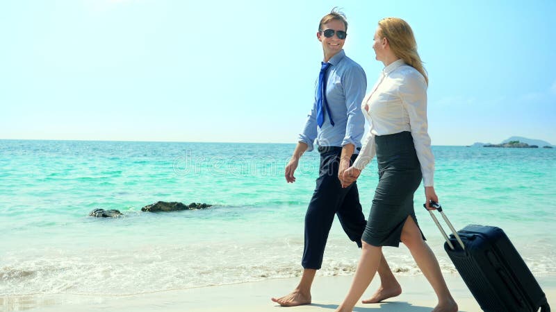 Businessman and business woman with a suitcase walking along the white sand beach on the island