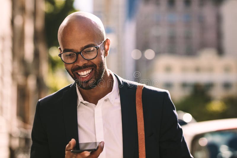 Smiling young african businessman commuting with a mobile phone while walking outdoors. Bald man in suit walking in the city and talking on cell phone. Smiling young african businessman commuting with a mobile phone while walking outdoors. Bald man in suit walking in the city and talking on cell phone.