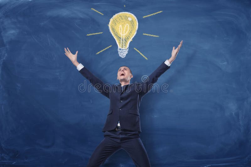 Businessman with hands raised in victory on blue blackboard background and a chalk drawing of a bulb right above him. stock photography