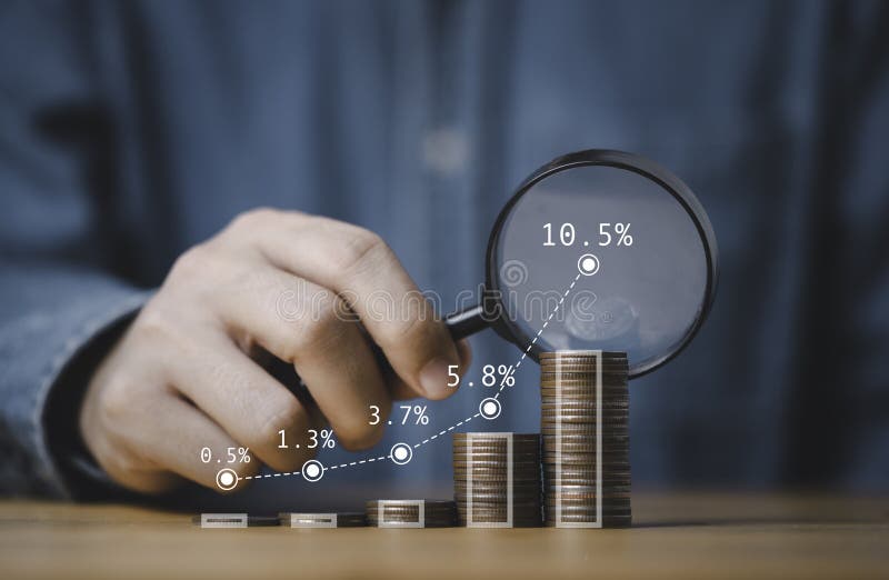 Businessman holding magnifier glass with coins stacking graph for finding the highest dividend or profit return from business investment and money saving concept.