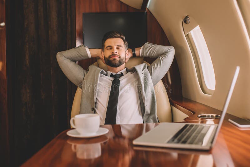 Businessman resting with hands behind head and closed eyes near laptop and cup of coffee in private plane