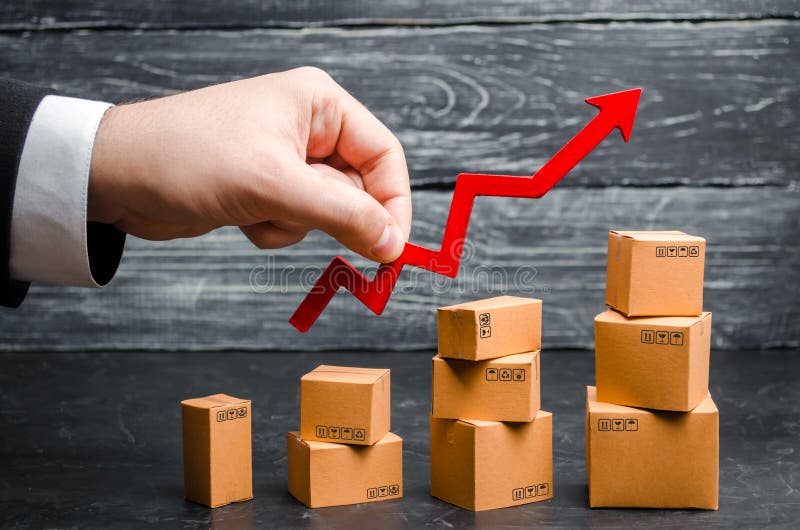 A businessman`s hand holds a red arrow up above cardboard boxes folded incrementally. Sales growth and increase in exports