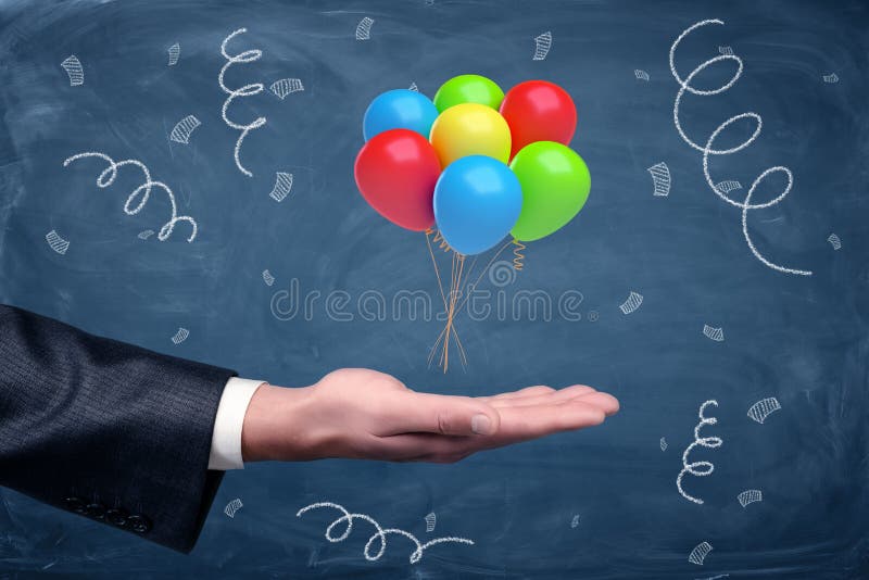 A businessman`s hand turned up with a set of colorful balloons tied together hovering above it on chalkboard background.